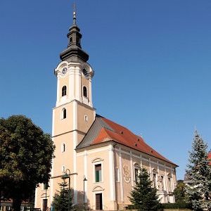 Eine alte Kirche mit einem hohen Turm und einem roten Dach steht auf einem Hügel mit Bäumen und Pflanzen darum herum. Die Kirche hat eine Uhr und einen Glockenturm.