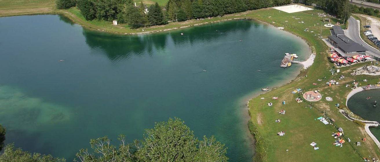 Ein Luftbild eines Badeortes am See mit einem Schwimmbecken, einem großen See und einer Berglandschaft im Hintergrund.