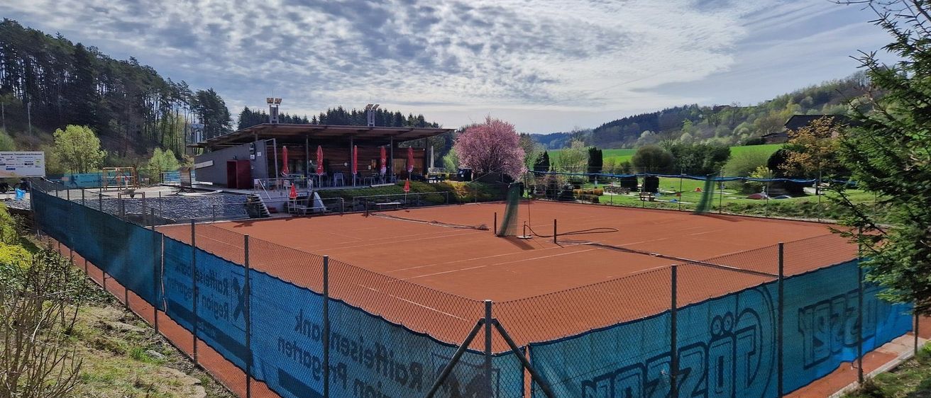 Ein Tennisplatz mit rotem Belag, umgeben von einem blauen Zaun, ist zu sehen. Im Hintergrund befindet sich ein Gebäude mit Dach und einigen Sonnenschirmen. Der Himmel ist blau mit weißen Wolken.