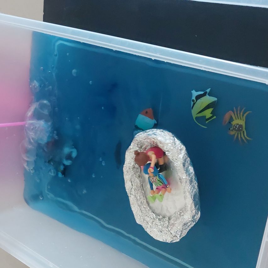 Ein Plastikbehälter mit blauem Wasser enthält ein silbernes Folienboot mit zwei Puppen darin, umgeben von Spielzeugfischen und Blasen.