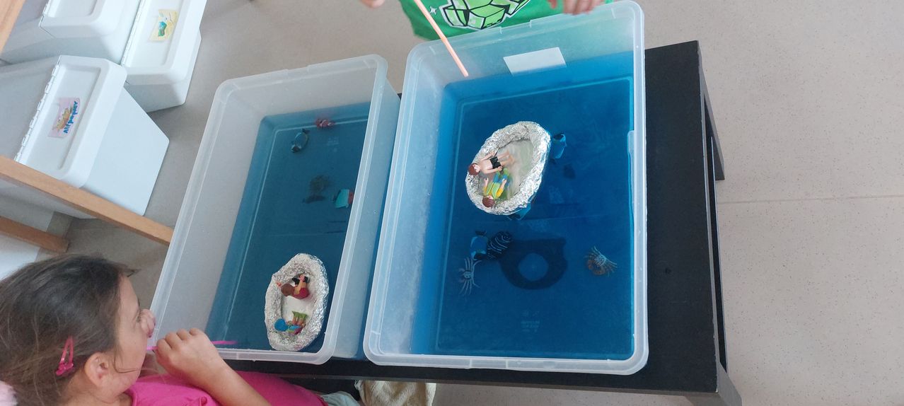 Zwei durchsichtige Plastikbehälter mit blauem Wasser, jeweils mit einem kleinen Boot mit Spielzeugfiguren darin. Ein Boot hat drei Figuren, das andere zwei.