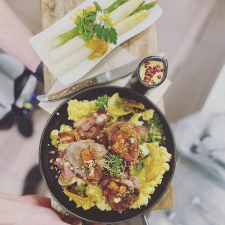 Ein Gericht mit Lammkoteletts auf einem Bett aus gelbem Reis und mit Grünzeug garniert wird von einer Person gehalten. Ein weißer Teller mit Spargel und einer weißen Blume befindet sich hinter dem Gericht.