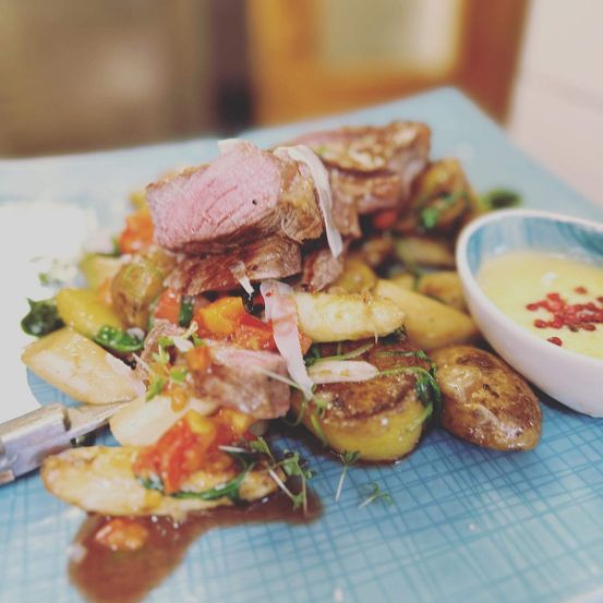 Ein Gericht mit Steak, Gemüse und Sauce auf einem blauen Teller, begleitet von einer kleinen Schüssel gelber Sauce.