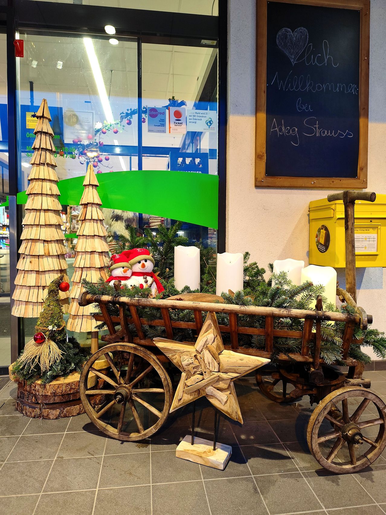 Ein hölzerner Weihnachtswagen mit Sterndekoration steht vor einem Schaufenster. Der Wagen ist mit Tannenzweigen, weißen Kerzen und zwei Schneemännern geschmückt.