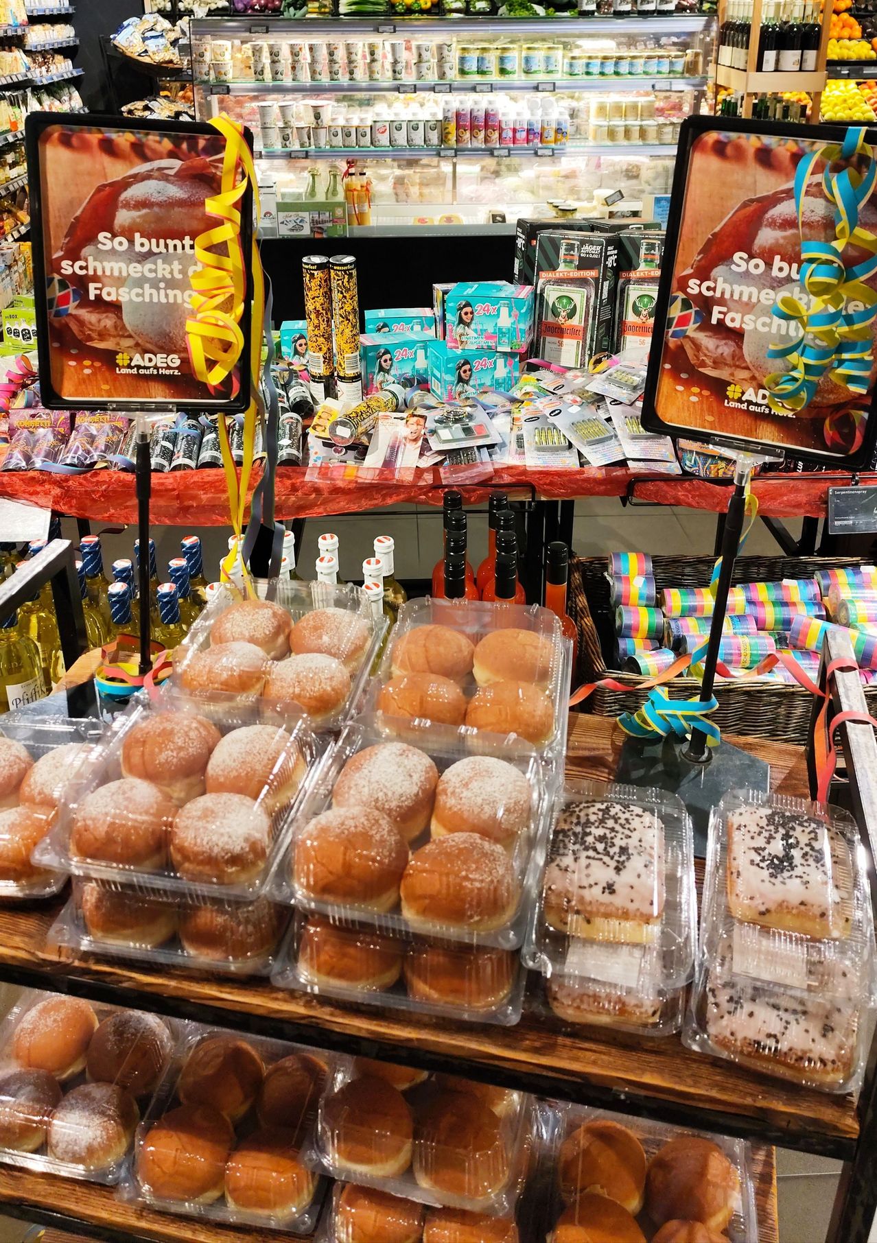 Eine Auslage von Gebäck und anderen Waren, mit zwei Schildern, die So bunt schmeckt Fasching lesen. Hinter dem Gebäck sind Flaschen angeordnet. Im Hintergrund befinden sich Produkte auf einem Tisch mit rotem Tuch.