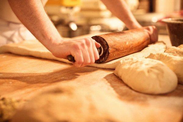 Eine Person rollt Teig auf einem Holztisch mit einem Nudelholz aus, wobei mehr Teig in der Nähe ist.