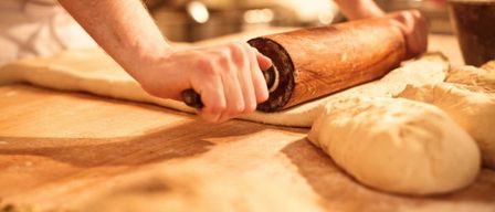 Eine Person rollt Teig auf einem Holztisch mit einem Nudelholz aus, wobei mehr Teig in der Nähe ist.