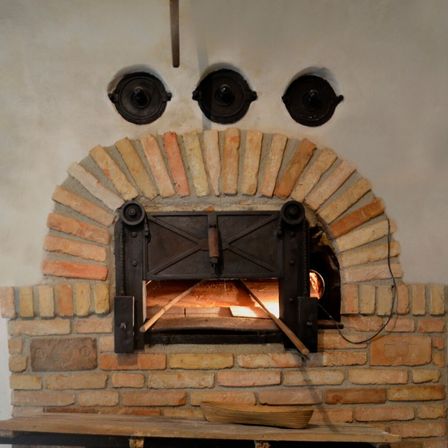 Ein alter Ziegelofen mit einer Metalltür und drei runden Metallplatten darüber. Ein Holztisch mit einem Korb steht davor.