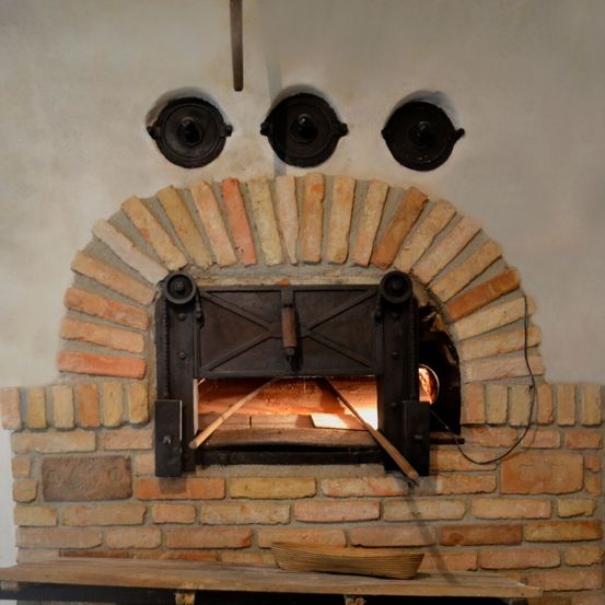 Ein alter Ziegelofen mit einer Metalltür und drei runden Metallplatten darüber. Ein Holztisch mit einem Korb steht davor.