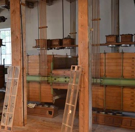 Industrieeinrichtung mit großen, gestapelten Holzkisten, die mit Metallstangen und Seilrollen verbunden sind, Holzleitern dazwischen und einem Fenster auf der linken Seite.