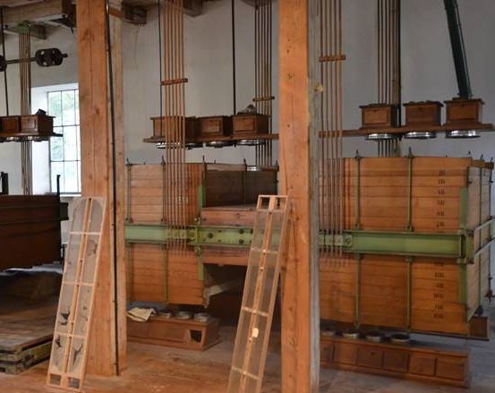 Industrieeinrichtung mit großen, gestapelten Holzkisten, die mit Metallstangen und Seilrollen verbunden sind, Holzleitern dazwischen und einem Fenster auf der linken Seite.