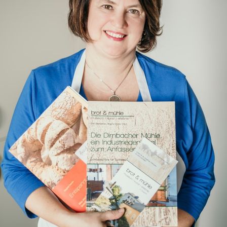 Eine lächelnde Frau in einem blauen Shirt und weißer Schürze hält Bücher und Broschüren, eines mit dem Titel 'brot & mühle'. Die Bücher zeigen Brot und Hausbilder.