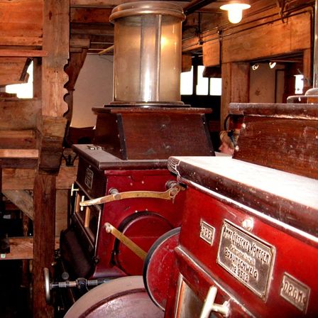 Eine alte rote Maschinerie mit Messingplatten und einer Glaslampe im Inneren eines Holzgebäudes. Eine Person steht hinter der Maschinerie.