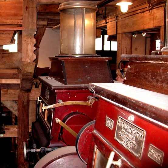 Eine alte rote Maschinerie mit Messingplatten und einer Glaslampe im Inneren eines Holzgebäudes. Eine Person steht hinter der Maschinerie.