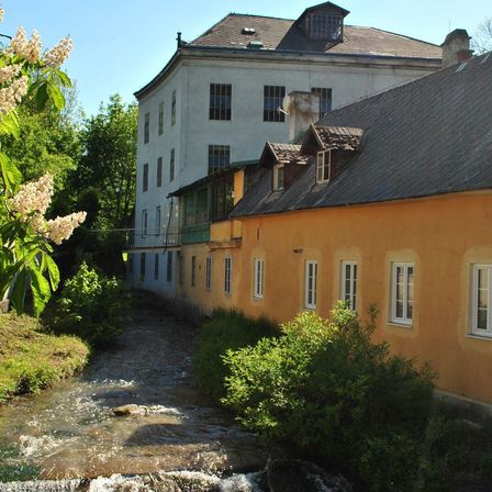 Ein klarer Bach fließt an einem gelben Gebäude mit grauem Dach vorbei, neben einem weißen Gebäude. Bäume und Pflanzen sind auf beiden Seiten sichtbar.