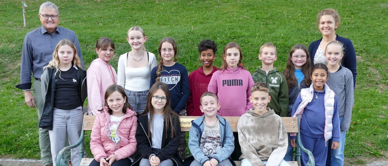 Eine Gruppe von Schülern und zwei Lehrern posiert für ein Foto im Park. Sie alle lächeln und stehen hinter einer Holzbank.