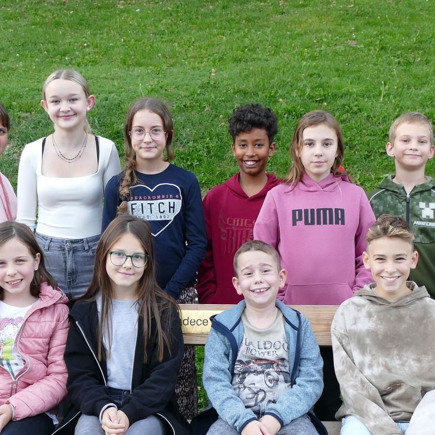 Eine Gruppe von Schülern und zwei Lehrern posiert für ein Foto im Park. Sie alle lächeln und stehen hinter einer Holzbank.