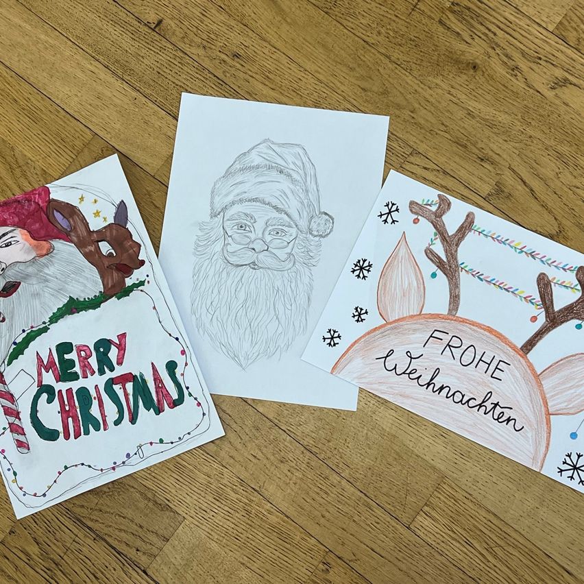 Drei Weihnachtskarten auf einer Holzoberfläche. Eine sagt Merry Christmas, eine andere ist eine Skizze von Santa und die letzte sagt Frohe Weihnachten.