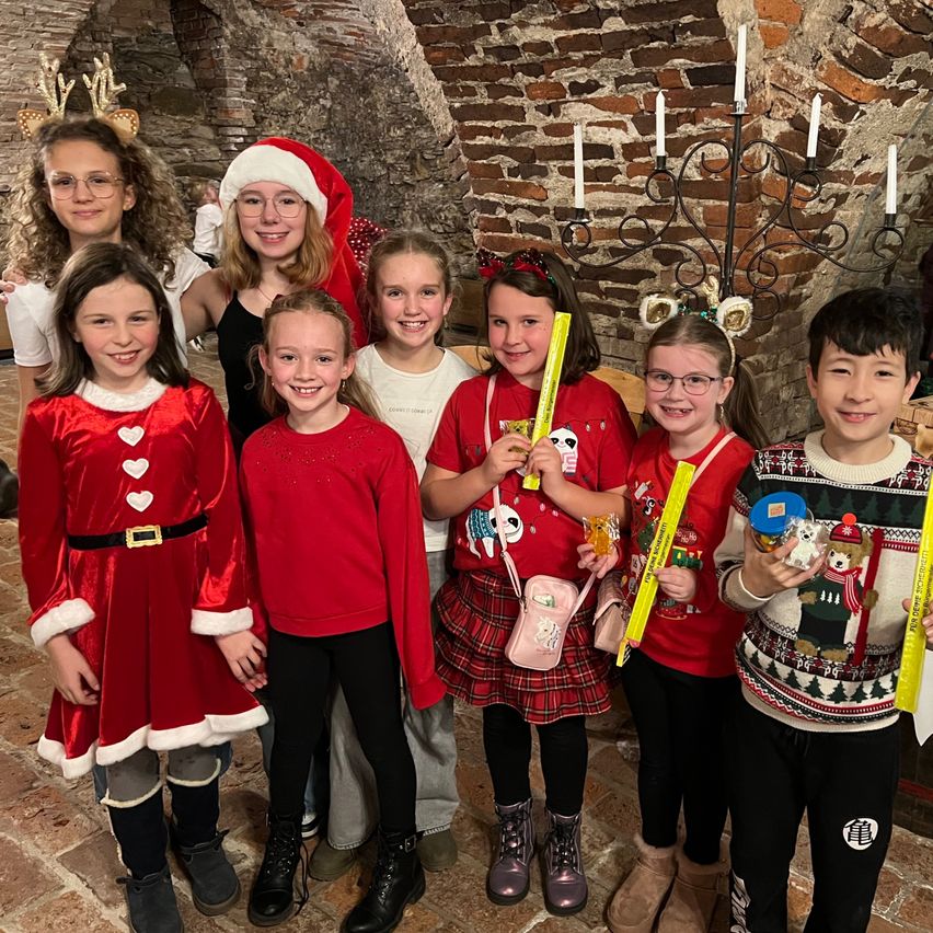 Eine Gruppe von Kindern in Weihnachtskleidung steht in einem Raum mit einer Ziegelmauer. Sie lächeln und halten verschiedene Gegenstände.