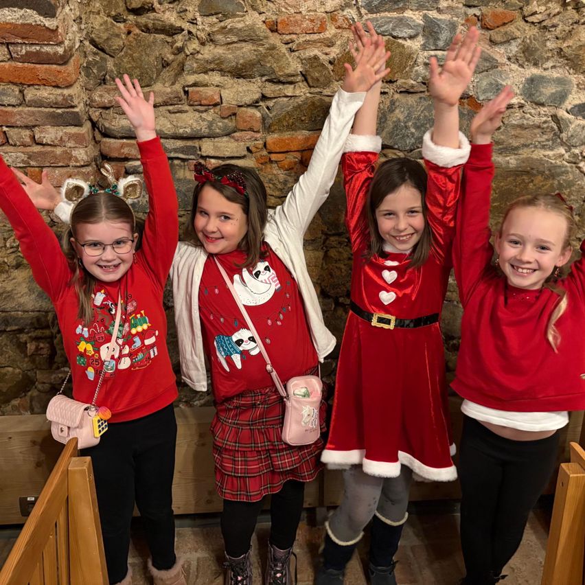 Fünf junge Mädchen in roten Festkleidern stehen zusammen vor einer Backsteinmauer, lächeln und heben ihre Hände.