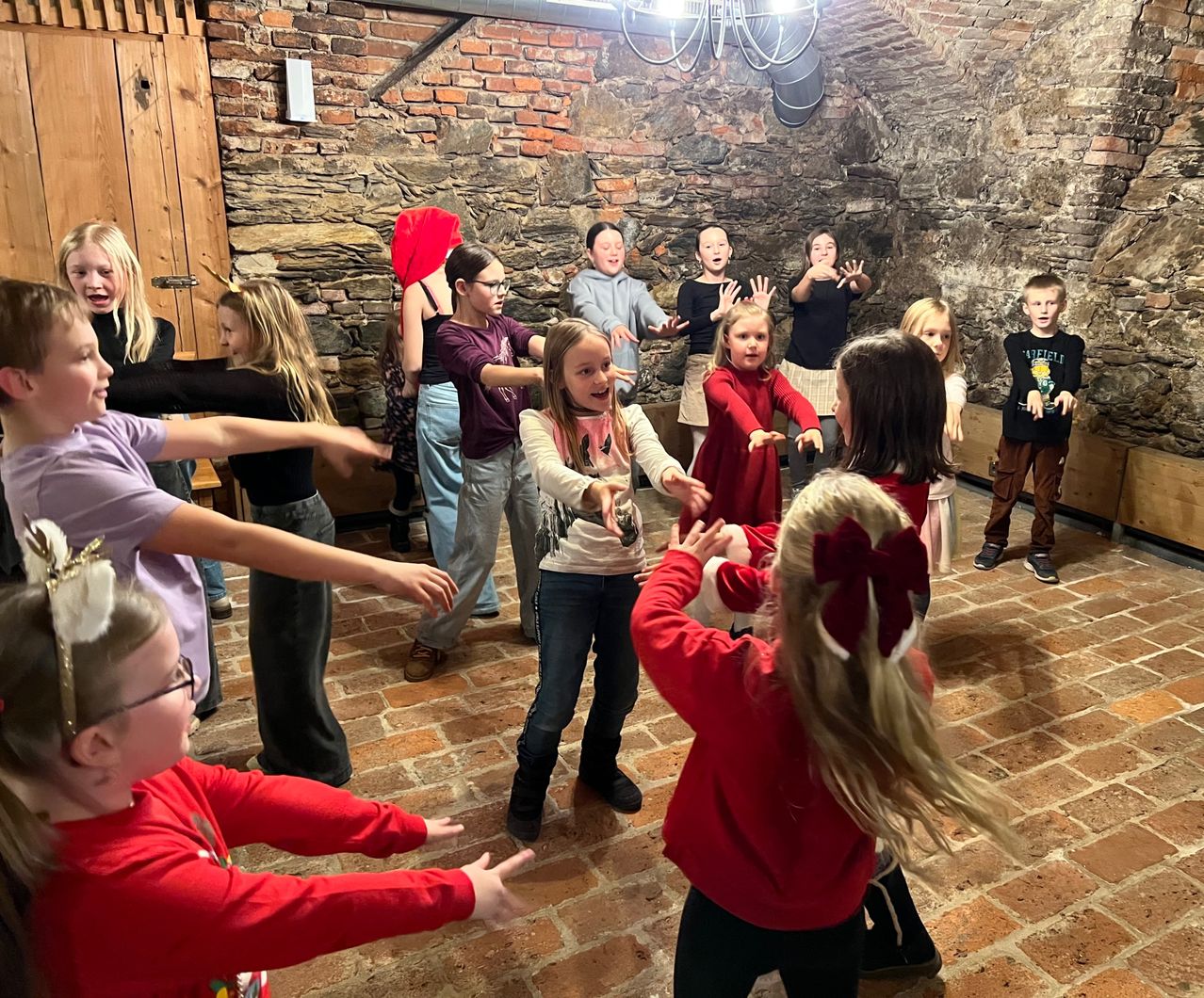 Eine Gruppe von Kindern in roten Hemden und Mützen tanzt in einem Raum mit Backsteinwänden. Einige tragen Brillen und Turnschuhe. Sie lächeln und machen Handgesten.