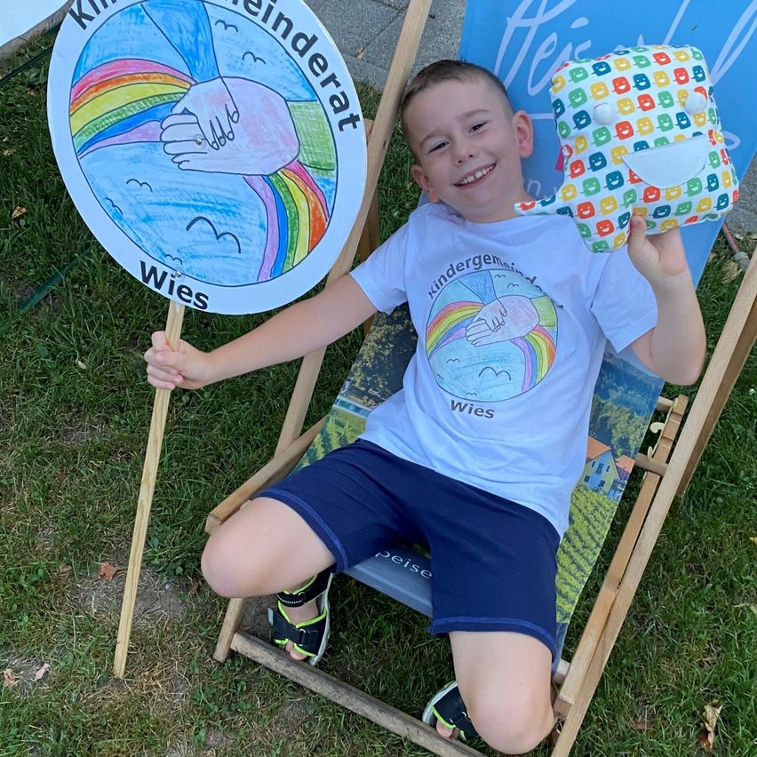 Ein junger Junge sitzt in einem Liegestuhl, lächelt und hält ein buntes Spielzeug und ein Schild mit einem Regenbogen und dem Wort 'Wies'.