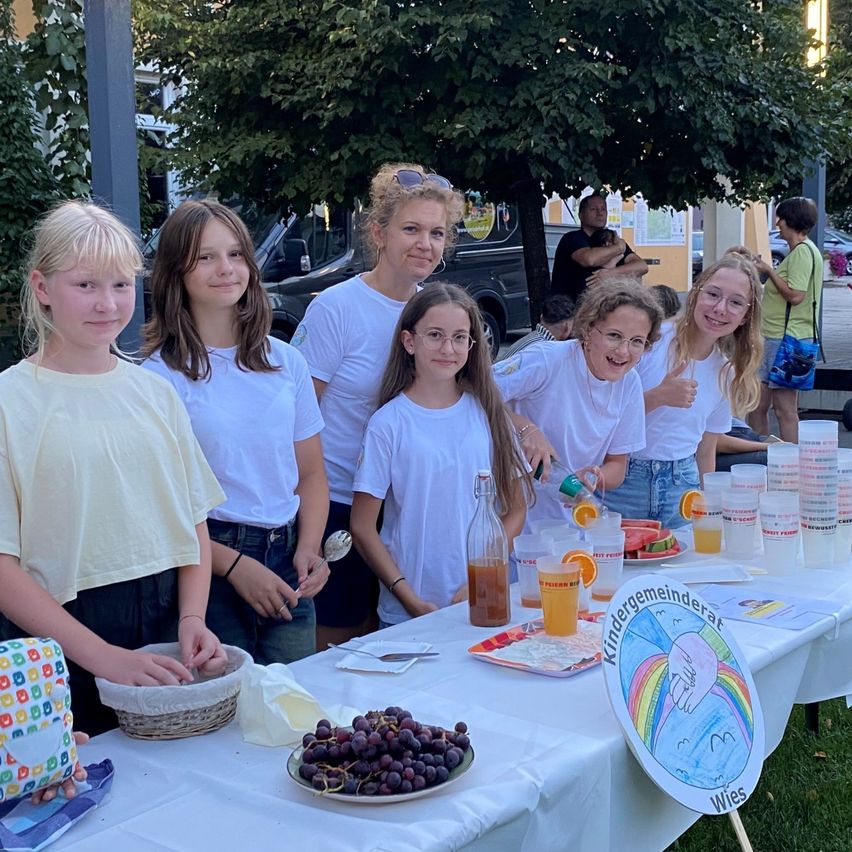 Eine Gruppe junger Mädchen und Frauen in weißen T-Shirts steht hinter einem Tisch mit Essen und Getränken, lächelt und posiert für ein Foto.