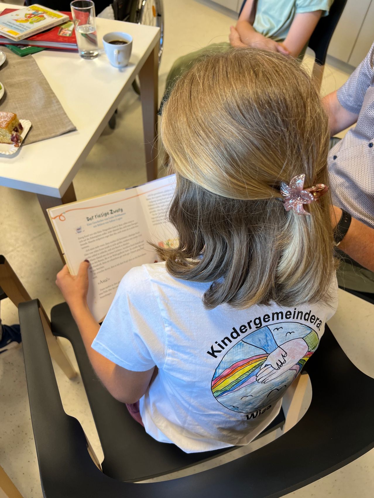 Ein junges Mädchen mit blonden Haaren liest ein Buch, während es auf einem Stuhl sitzt. Sie trägt ein weißes T-Shirt mit farbenfrohen Handzeichnungen. Eine Person steht hinter ihr. Im Hintergrund ist ein Tisch mit einem Teller Kuchen zu sehen.