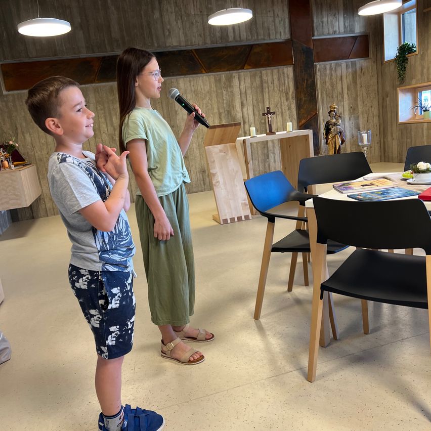 Ein junges Mädchen hält ein Mikrofon in der Hand und steht neben einem Jungen in einem Raum. Sie schauen beide nach oben. Hinter ihnen befindet sich ein Tisch mit Stühlen und eine Holzwand.