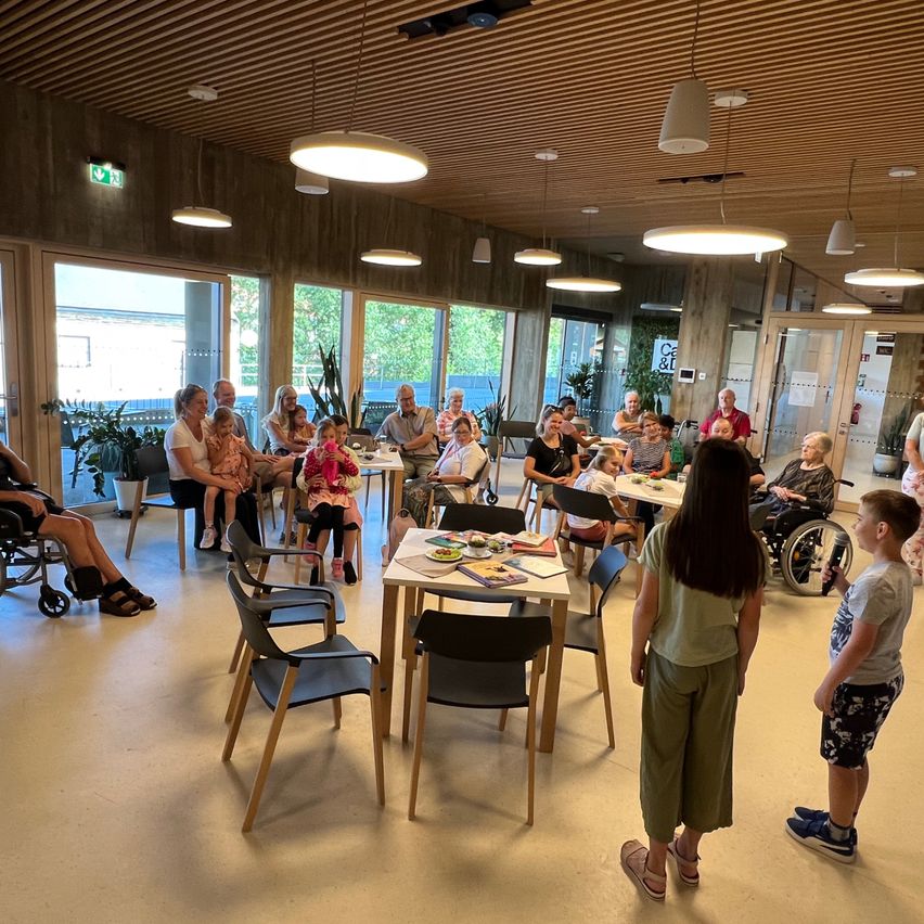 Eine Gruppe von Erwachsenen und Kindern sitzt an Tischen in einem gut beleuchteten Raum, möglicherweise einem Gemeindezentrum. Topfpflanzen und Lampen hängen von der Decke. Eine Frau und ein Junge stehen vor ihnen und unterhalten sich mit der Gruppe.