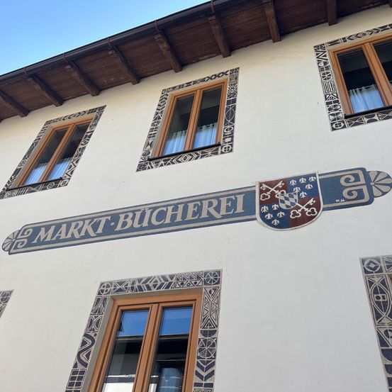 Ein Gebäude mit einem Schild, das 'Markt-Bucherei' besagt, einem Wappenschild und Fenstern mit Vorhängen. Das Gebäude hat eine weiße Wand und ein Holzdach.