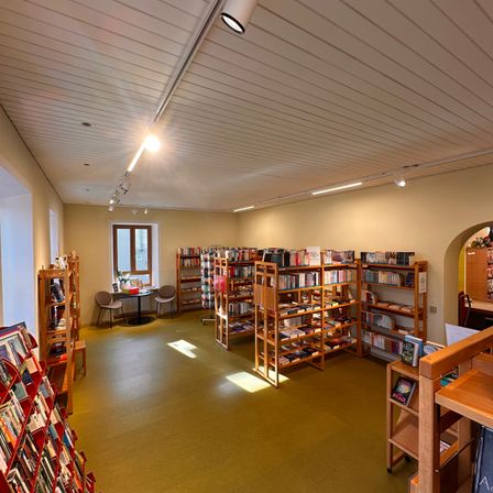 Ein Innenansicht einer Bibliothek mit Bücherregalen aus Holz, einem kleinen Tisch mit Stühlen und Sonnenlicht, das durch ein Fenster strömt.
