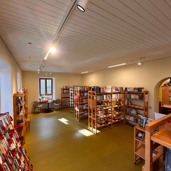 Ein Innenansicht einer Bibliothek mit Bücherregalen aus Holz, einem kleinen Tisch mit Stühlen und Sonnenlicht, das durch ein Fenster strömt.
