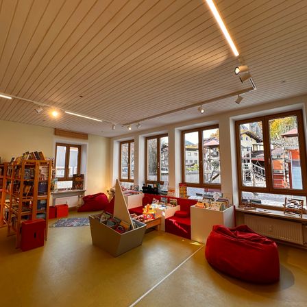 Ein heller Raum mit gelbem Boden, Holzdecke und großen Fenstern. Es gibt Sitzsäcke, einen Bücherregal, einen Tisch mit Büchern und einen Heizkörper. Draußen sind Gebäude und Bäume durch die Fenster zu sehen.
