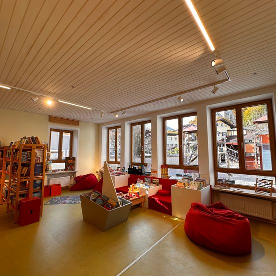 Ein heller Raum mit gelbem Boden, Holzdecke und großen Fenstern. Es gibt Sitzsäcke, einen Bücherregal, einen Tisch mit Büchern und einen Heizkörper. Draußen sind Gebäude und Bäume durch die Fenster zu sehen.