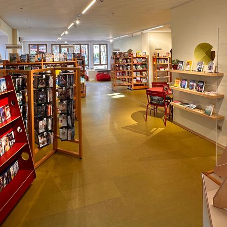 Ein Innenraum eines Buchladens mit Holzregalen und Büchern, mit heller Beleuchtung und gelbem Boden. Die Regale sind mit verschiedenen Büchern gefüllt, und es gibt Tische und Stühle im Bereich.