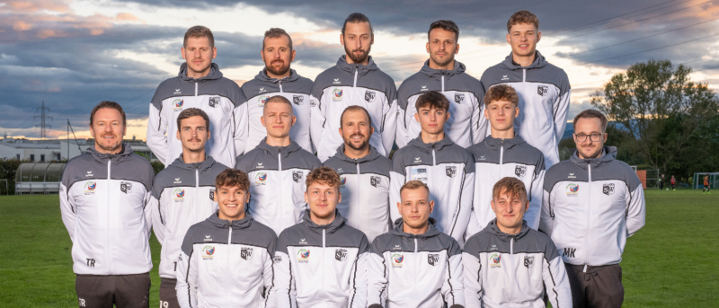 Eine Gruppe von Fußballspielern posiert für ein Mannschaftsfoto auf einem Rasenfeld bei bewölktem Himmel. Sie tragen weiße und graue Trikots mit Nummern und Logos. Sie sitzen auf einer Bank, einige stehen dahinter.