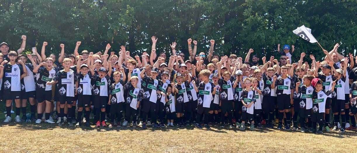 Eine Gruppe junger Spieler und Trainer feiert gemeinsam an einem sonnigen Tag. Sie tragen alle passende Uniformen mit grünen Logos. Sie posieren für ein Foto mit erhobenen Händen. Dahinter stehen Bäume.