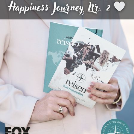Eine Person hält zwei Bücher mit dem Titel 'reisen mit Freunden'. Das oberste Buch hat eine Weltkarte auf dem Cover. Die Person trägt einen weißen Blazer, Ringe und eine Armbanduhr. Das Bild ist Teil der 'Happiness Journey Nr. 2'-Serie.