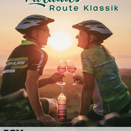 Zwei Radfahrer in Fahrradausrüstung genießen Wein auf einem sonnenuntergangsbeleuchteten Hügel. Eine Flasche Wein steht zwischen ihnen.