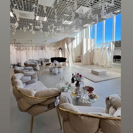 Ein modernes Brautmodengeschäft mit weißen Wänden, großen Fenstern und eleganten Möbeln. Ausgestellt sind mehrere Brautkleider und eine Reihe eleganter Sitzgelegenheiten.