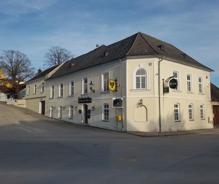 Ein großes weißes Gebäude mit grauem Dach befindet sich an einer Ecke mit mehreren Fenstern und Schildern, unter einem klaren Himmel.