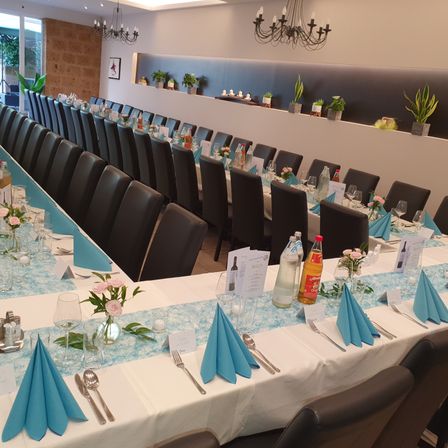 Ein großer Speisesaal ist für ein Bankett vorbereitet mit blau-weißen Tischdecken, Blumensträußen und einer langen Reihe von Stühlen. Flaschen, Gläser und Besteck sind ordentlich arrangiert. Die Wand hat Regale mit Topfpflanzen, und Kronleuchter hängen von der Decke.