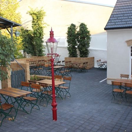 Ein Außenbereich eines Restaurants mit Holztischen und Stühlen, einem roten Laternenpfahl und Pflanzen. Das Gebäude hat ein Fenster und ein Ziegeldach.