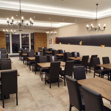 Ein Restaurant mit modernem Interieur, Holztischen, schwarzen Stühlen und Kronleuchtern. Der Raum hat beige Wände, eine offene Tür und Dekorationen an der Wand.