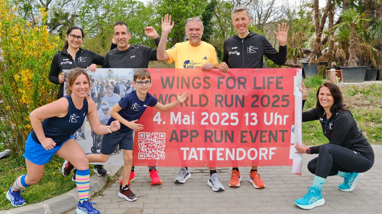 Fünf Personen posieren hinter einem Banner für den Wings for Life World Run 2025. Das Banner hat einen QR-Code, ein Bild eines Läufers und das Datum und die Uhrzeit der Veranstaltung. Die Person in der Mitte ist ein junger Junge mit Brille. Sie tragen alle Turnschuhe.