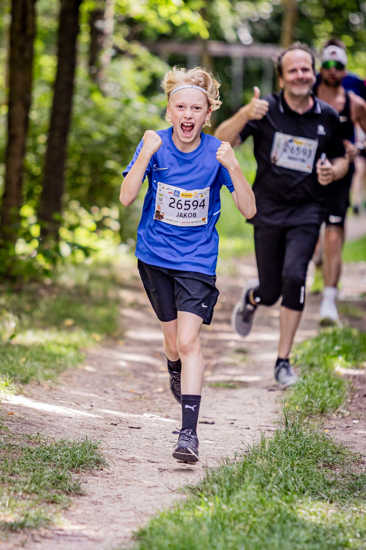Ein Junge läuft in einem Marathon mit offenem Mund. Er trägt ein blaues Shirt mit der Nummer 26594. Ein anderer Läufer folgt ihm.