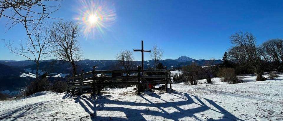 Ein Kreuz auf einem Hügel im Schnee, ein Holzzaun und ein blauer Himmel mit einem strahlenden Sonnenschein.