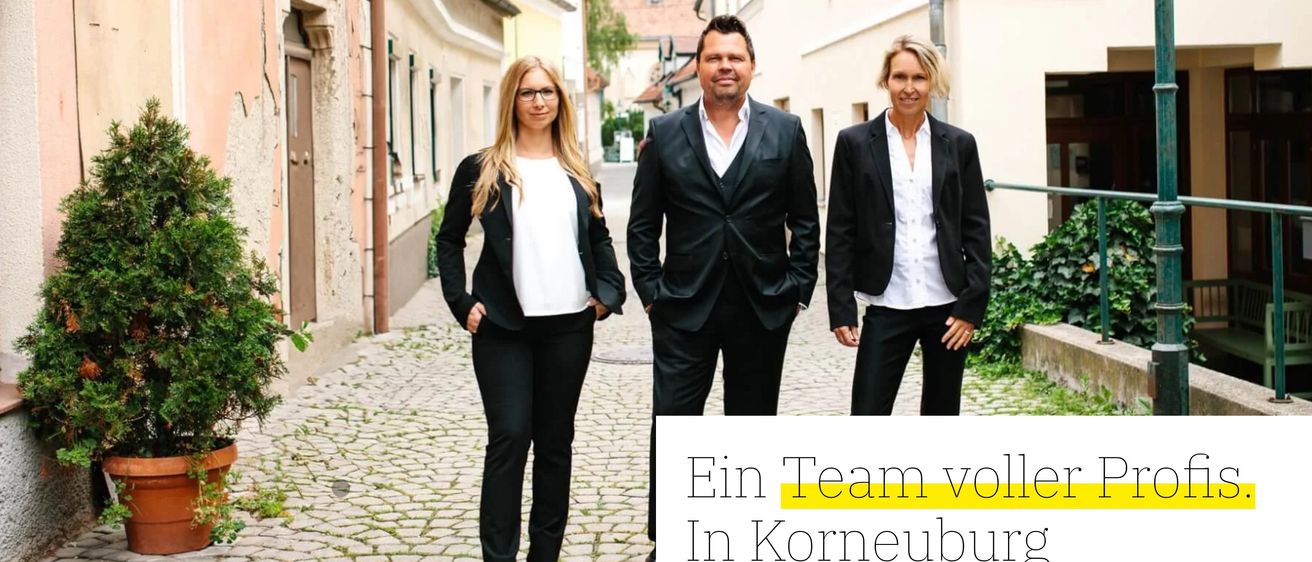 Drei Personen, zwei Frauen und ein Mann, stehen zusammen auf einer Kopfsteinpflasterstraße vor Gebäuden und lächeln für ein Foto. Das Bild ist mit 'Ein Team von in Korneuburg' beschriftet.