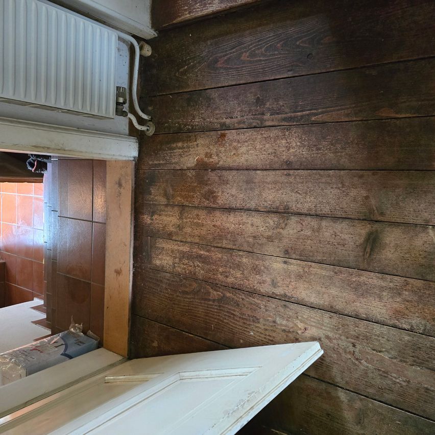 Eine Holzwand, wahrscheinlich Teil eines Hauses, weist sichtbare Abnutzungserscheinungen auf. Die Wand besteht aus Holzbrettern, von denen einige Flecken und Markierungen aufweisen. Unten im Bild befindet sich eine weiße Tür mit weißem Rahmen.